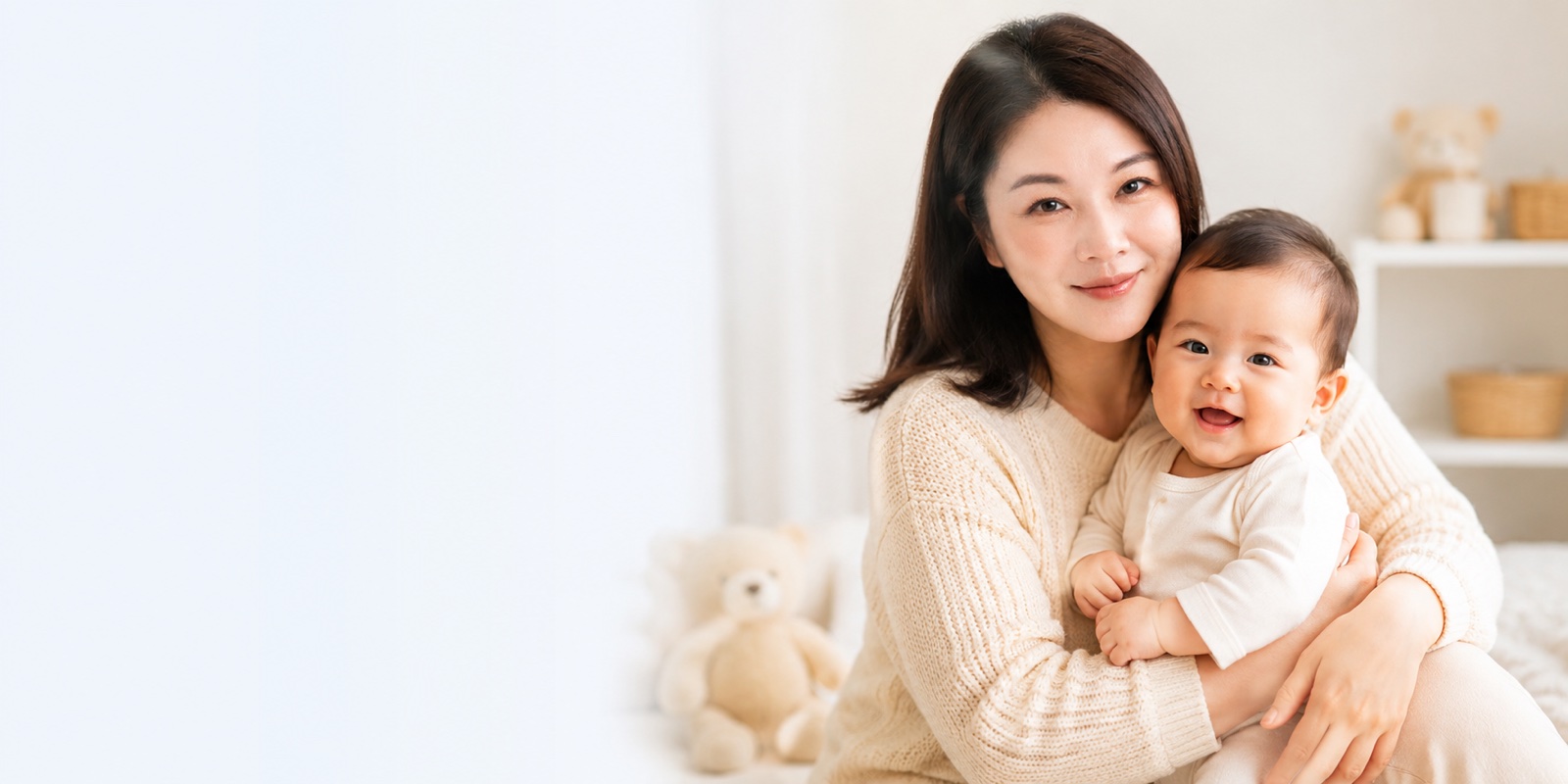 Asian mother gently holding her smiling baby on a soft bed with a teddy bear in the background, warm natural light