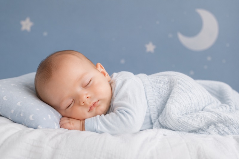 Sleeping baby in a blue onesie against a navy nursery wall painted with moon and stars